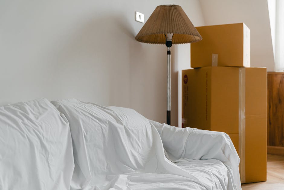Inside a home during a furniture transport process, a white couch covered with a protective white sheet is positioned on the left side of the image. Behind it, in the corner near a wooden wall, there are three cardboard boxes stacked, varying in size, with the smallest on top. To the right of the boxes, a tall floor lamp with a brown pleated lampshade is visible. The environment is well-lit, with natural light coming from a window on the right, which is partially out of frame. The scene appears to be part of a home relocation, with the packing and loading process underway, supported by removals company Removals Earls Court, as evident from the professional packing materials and careful arrangement of furniture and boxes inside the property.