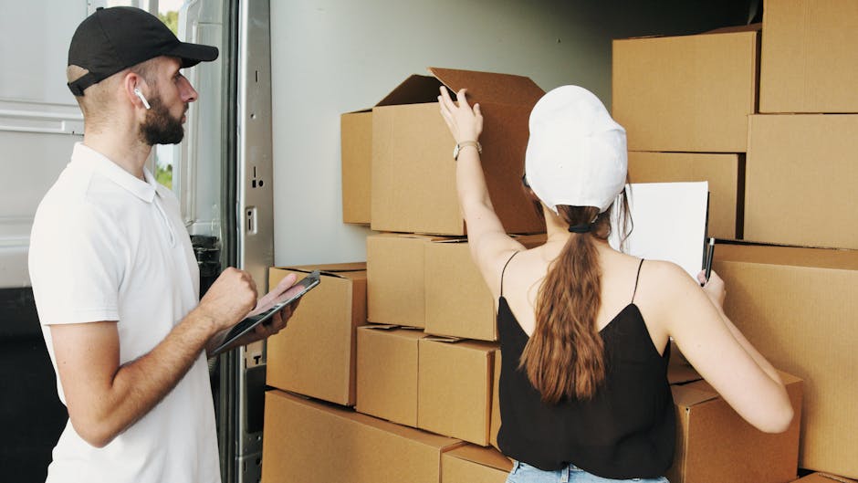 A man with a beard and dark cap dressed in a white polo shirt is inside a property, standing next to a stack of sealed cardboard boxes of various sizes. He is holding a clipboard and writing, focusing on the packing and organization process. The boxes are arranged on a flat surface, possibly a table or the floor, and some are stacked against a wall, indicating preparations for home relocation or furniture transport. The environment is well-lit with natural light, and the scene captures the meticulous planning associated with professional removals, as seen with the attention to packing materials and the presence of the boxes. This visual aligns with the services offered by Removals Earls Court, emphasizing careful packing and moving logistics for house removals.